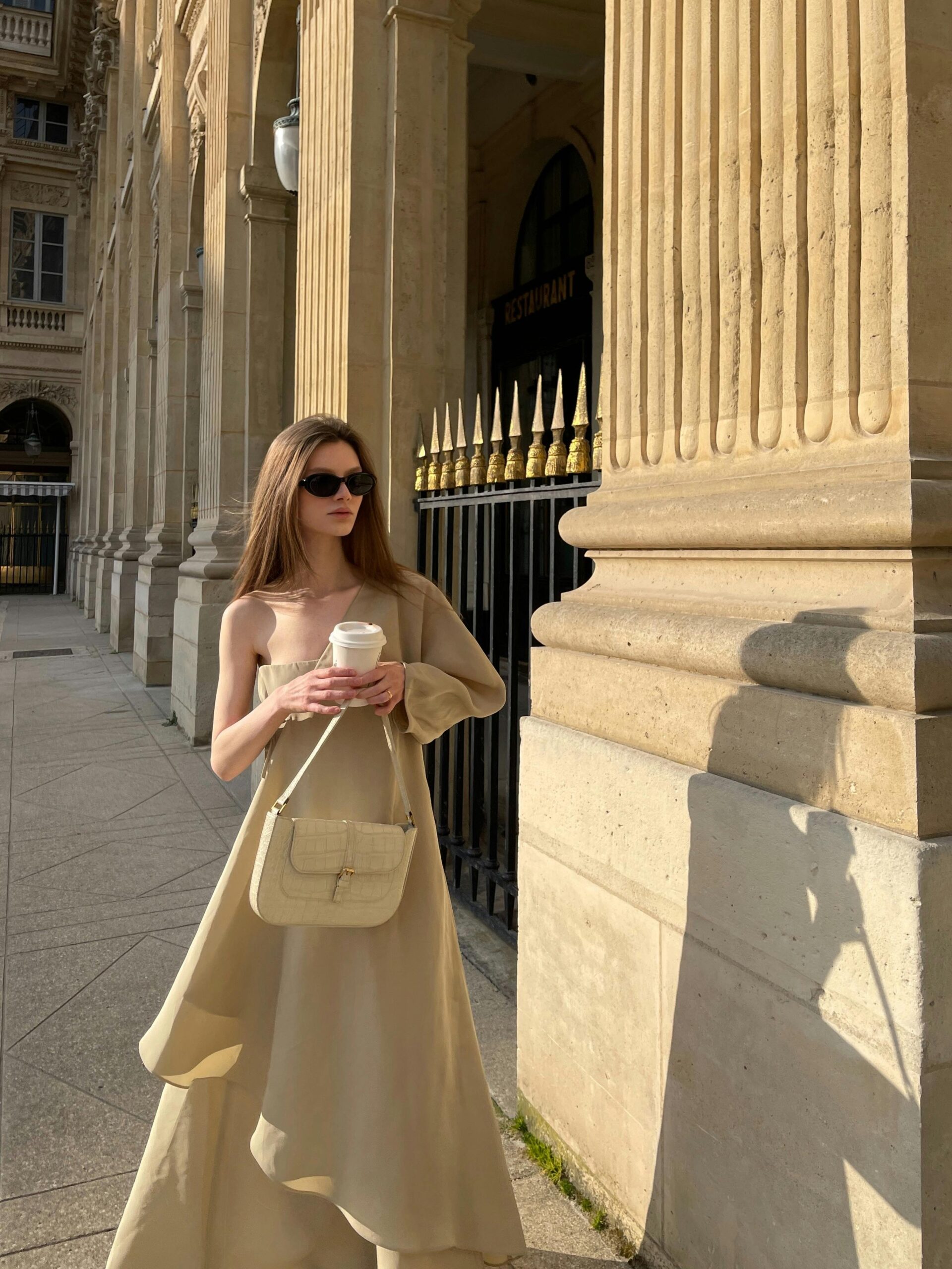 Elegant woman in a flowing beige dress walking along a sunlit architectural pathway, holding a coffee cup.